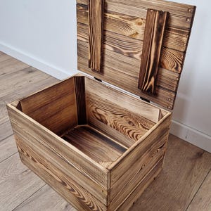 May include: An open wooden storage chest with a hinged lid. The chest is made of light brown wood with darker brown accents from the wood grain. The lid is propped open, revealing the empty interior. The chest is sitting on a wooden floor.