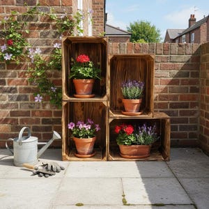 Puede incluir: Cuatro cajas de madera apiladas para crear una exhibición de plantas en macetas. Cada caja contiene una maceta de terracota con flores coloridas, incluyendo geranios rojos, lavanda y petunias rosas. También se ve una regadera de metal, una paleta y guantes.