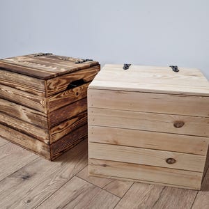 May include: Two wooden storage boxes with hinged lids. One box is stained a dark brown, and the other is natural wood. Both boxes are square and made of horizontal wooden planks. The boxes are on a wooden floor.