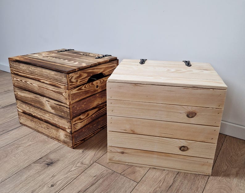 May include: Two wooden storage boxes with hinged lids. One box is stained a dark brown, and the other is natural wood. Both boxes are square and made of horizontal wooden planks. The boxes are on a wooden floor.