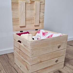 May include: A natural wood storage chest with a hinged lid, partially open. The chest is filled with folded blankets and other items. The chest is made of light-colored wood and has a rustic appearance. The lid is attached with hinges.