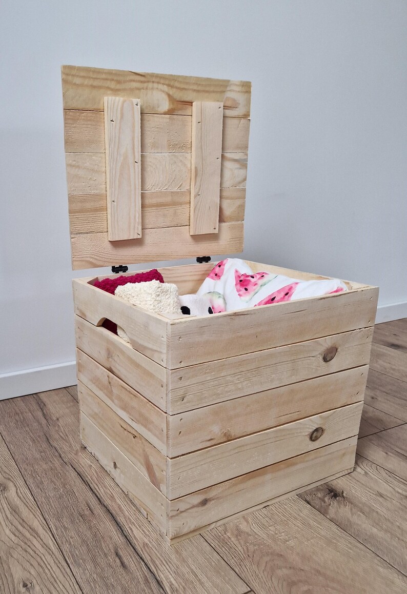 May include: A natural wood storage chest with a hinged lid, partially open. The chest is filled with folded blankets and other items. The chest is made of light-colored wood and has a rustic appearance. The lid is attached with hinges.