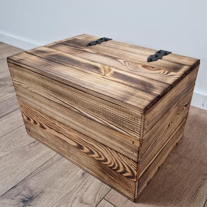 May include: A rustic wooden storage box with a hinged lid. The box is constructed from light brown planks with dark brown burn marks, and features black metal hinges. The wood grain is visible, adding to the box's natural aesthetic.