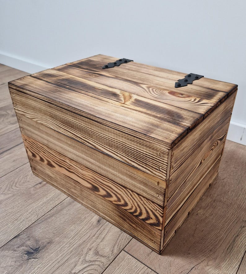 May include: A rustic wooden storage box with a hinged lid. The box is constructed from light brown planks with dark brown burn marks, and features black metal hinges. The wood grain is visible, adding to the box's natural aesthetic.
