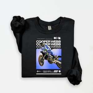 May include: Black sweatshirt with a graphic of a motocross rider in mid-air, with the text "COOPER WEBB" repeated three times. The graphic also includes the text "MONSTER ENERGY" and "YAMAHA STAR RACING".