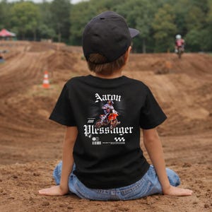 May include: A black t-shirt featuring a graphic print of a motocross rider on a motorcycle with the text Aaron Plessinger, Red Bull Factory Racing, and AMA SX Champion. The shirt is worn with a plain black baseball cap and blue denim jeans.