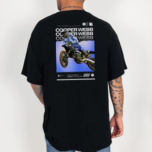 May include: Black t-shirt with a graphic print of a motocross rider on a blue background. The text "COOPER WEBB" is repeated three times on the shirt. The text "MONSTER ENERGY" and "YAMALUBE STAR RACING" are also printed on the shirt.