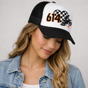 May include: Black and white trucker hat with a white front panel featuring the number "614" in orange, a motorcycle graphic, and a checkered heart design. The hat has a black mesh back and a curved black brim.