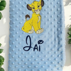 Puede incluir: Una manta para bebé azul claro con una superficie texturizada y una imagen bordada de Simba de El Rey León. El nombre "Jai" está bordado en azul oscuro debajo de la imagen.