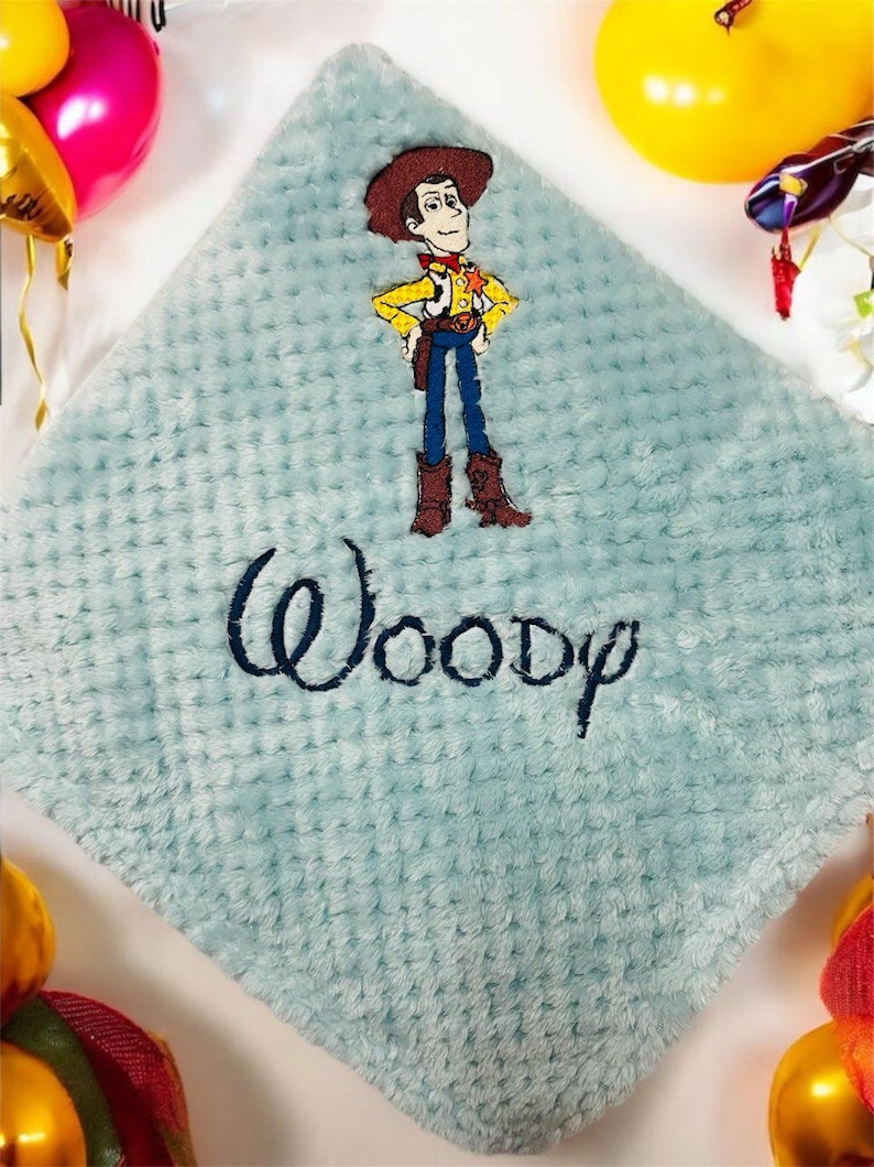 Puede incluir: Una manta de beb&eacute; azul claro y texturizada con una imagen bordada del personaje Woody de Toy Story. El nombre "Woody" est&aacute; bordado en escritura azul marino. La manta es de forma cuadrada.