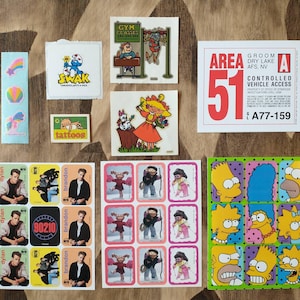 May include: A collection of colorful stickers featuring various cartoon characters, including a blue character with a red hat, a yellow character with a red hat, and a yellow character with a blue hat. The stickers also include text such as "SWAK", "tattoos", "GYM FLAGSES", "AREA 51", and "A77-159".