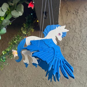 Könnte beinhalten: Ein skurriles Mobile mit einem weißen und blauen Pegasus mit ausgebreiteten Flügeln. Der Pegasus hängt an Schnüren, darunter eine kleine weiße Wolke. Das Mobile ist zum Aufhängen konzipiert und verleiht jedem Raum einen Hauch von Fantasie.