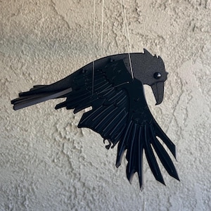 Puede incluir: Un móvil con forma de cuervo negro suspendido de un gancho. El cuervo está elaborado con diseños detallados de alas y plumas. El fondo es una pared texturizada de color claro, con vegetación y luces de hadas en la esquina superior izquierda.