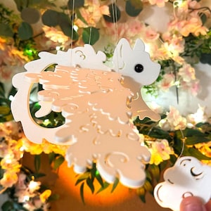 Peut inclure: Mobile licorne blanche découpée au laser avec un nuage. La licorne a une crinière et une queue fluides, suspendues par de fins fils. Le fond présente des fleurs roses et des guirlandes lumineuses, créant un thème décoratif fantaisiste.