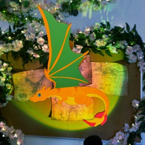 Könnte beinhalten: Hängende Drachendekoration mit orangefarbenem Körper, grün-orangen Flügeln und einem rot-gelben Schwanz. Der Drache hängt vor einem Hintergrund aus alten Karten und Blumengestecken. Der Drache hat ein cartoonartiges Design.