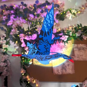 May include: A blue and black decorative wooden mobile shaped like a flying creature, with red accents on the wings. The mobile is suspended in front of a floral background with pink and white blossoms and fairy lights.