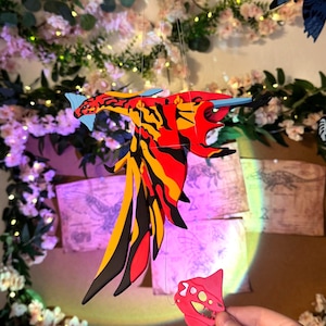 Op de afbeelding: Een kleurrijke, handgemaakte mobiel met een gestileerd, gevleugeld wezen met rode, gele en zwarte patronen. De mobiel hangt tegen een achtergrond van bloemversieringen en feeërieke lichtjes, met extra soortgelijke figuren op de achtergrond.