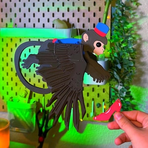 May include: A hanging ornament featuring a brown monkey with a blue and red hat, wearing a blue vest, and attached to large dark brown wings. A thin string connects the monkey to a small, bright red high-heeled shoe hanging below it.
