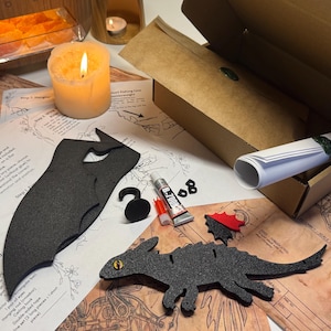 May include: A craft kit with dragon-themed cutouts, glue, and instructions. The kit includes a black dragon silhouette, a red and black wing piece, and a hook. A lit candle and a box with a rolled-up scroll are also visible.