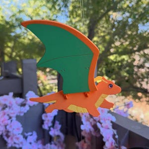 Può includere: Una campanella a vento a forma di drago arancione con ali verdi e dettagli gialli. Il drago è sospeso a mezz'aria, con uno sfondo sfocato di alberi e fiori. Un oggetto decorativo per un giardino o un patio.