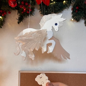 Pode incluir: Um mobile de Pégaso branco com asas translúcidas e olhos azuis pendurado em frente a uma grinalda de Natal. Um ímã branco em forma de nuvem é segurado em primeiro plano. O fundo inclui um quadro de avisos.