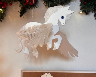 May include: A white Pegasus mobile with translucent wings and blue eyes hangs in front of a Christmas garland. A white cloud-shaped magnet is held in the foreground. The background includes a bulletin board.
