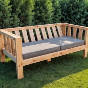 May include: A wooden outdoor bench with a slatted back and armrests. The bench has two gray cushions and is set on a green lawn. The wood has a natural finish.