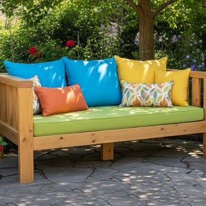 May include: A wooden outdoor daybed with a lime green cushion and several colorful pillows. The pillows are in shades of blue, orange, yellow, and patterned with leaf designs. The daybed is set on a stone patio in a garden setting.