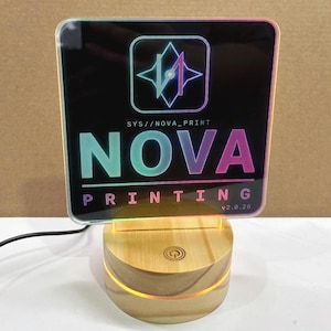 Może przedstawiać: Kwadratowy szyld akrylowy z czarnym tłem i podświetlaną drewnianą podstawą. Szyld zawiera geometryczne logo, tekst SYS//NOVA_PRINT, słowo NOVA dużymi literami z gradientem, słowo PRINTING oraz tekst v2.0.26 w rogu.