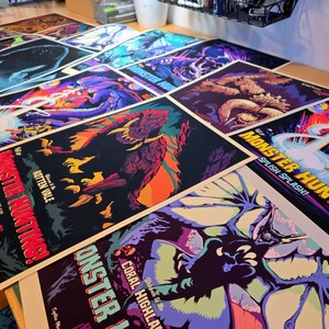 May include: A collection of colorful posters featuring monster illustrations and text. The posters have a variety of designs, including images of creatures and text that reads "MONSTER HUNTING!" and "SPLISH SPLASH!" The posters are on a wooden surface.