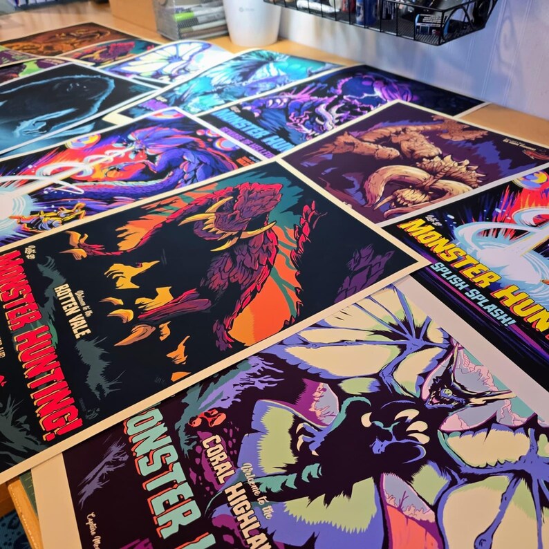 May include: A collection of colorful posters featuring monster illustrations and text. The posters have a variety of designs, including images of creatures and text that reads "MONSTER HUNTING!" and "SPLISH SPLASH!" The posters are on a wooden surface.
