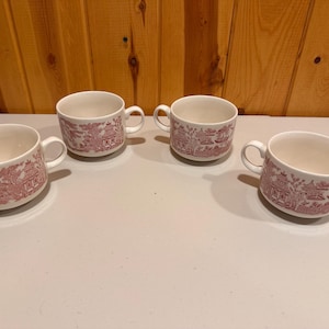 May include: Four white ceramic mugs with a red Willow pattern design. Each mug has a handle and a wide opening. The mugs are arranged on a light-colored surface, with a wooden background.