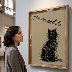 Könnte beinhalten: Ein gerahmtes Kunstwerk mit einer schwarzen Kritzelkatze auf beigem Hintergrund. Der Text "you, me, and the" steht über der Katze. Das Kunstwerk wird in einer Galerie ausgestellt.