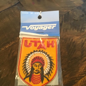 May include: A yellow and red embroidered patch with the word "UTAH" in red letters and a Native American chief wearing a headdress. The patch is on a white background with a blue and white Voyager label.