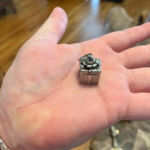 May include: A small, silver-colored charm in the shape of a gift box, with a bow on top. The charm is held in a person's open palm. The charm has a textured surface and is detailed with lines and a bow.