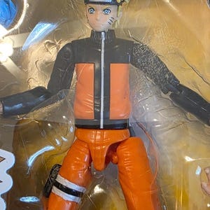 Op de afbeelding: Een Naruto actiefiguur in een doorzichtige plastic verpakking. De figuur heeft stekelig geel haar, blauwe ogen en een zwart-oranje outfit. De figuur staat in een dynamische pose met uitgestrekte armen.