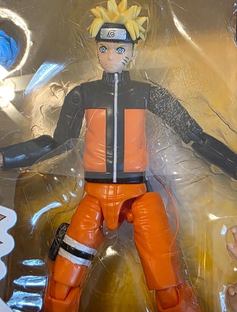 Op de afbeelding: Een Naruto actiefiguur in een doorzichtige plastic verpakking. De figuur heeft stekelig geel haar, blauwe ogen en een zwart-oranje outfit. De figuur staat in een dynamische pose met uitgestrekte armen.