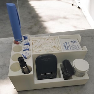 May include: White plastic organizer with compartments for medical supplies. It includes a blood glucose meter, lancets, alcohol prep pads, and a pen. The meter is black with a screen that says "Accu-Chek Guide".