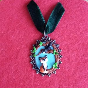 Peut inclure: Un collier avec un pendentif ovale en argent représentant un corbeau noir et un renard roux sur une branche avec des feuilles vertes. Le pendentif est suspendu à un ruban de velours vert.