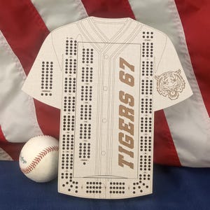 May include: A light-colored wooden cribbage board shaped like a baseball jersey, featuring the text TIGERS 67, a tiger head graphic, and numbered scoring holes. A white baseball with red stitching is positioned next to the board.