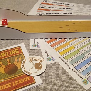 May include: A tabletop bowling game set with a wooden lane and a red die. The set includes score sheets, a bowling-themed card, and a "Perfect Game" sticker. The game is labeled "Dice League Bowling".