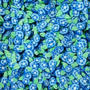 Könnte beinhalten: Nahaufnahme von Polymer-Ton-Blaubeerperlen. Die Perlen sind überwiegend blau und weiß und weisen ein kreisförmiges Muster auf. Grüne Blattakzente sind in der Anordnung verstreut und erzeugen ein natürliches Aussehen.