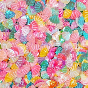 Puede incluir: Un vibrante surtido de adornos iridiscentes en forma de concha marina en una variedad de colores, incluyendo rosa, amarillo, verde y azul. Estas piezas decorativas están dispersas, creando una superficie colorida y texturizada.