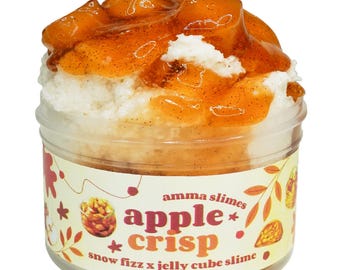 May include: A jar of "apple crisp" slime. The slime has a white base topped with a layer of clear, amber-colored jelly. The jar's label features the words "apple crisp" in orange and red, with the words "snow fizz x jelly cube slime" below.