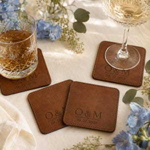 May include: Four square brown leather coasters with rounded corners, each engraved with the letters O & M, a small heart symbol, and the date 06.22.2026. The coasters feature visible stitching along the edges and a smooth, matte finish.
