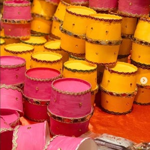 Puede incluir: Una pila de coloridos recipientes cilíndricos en tonos rosa y amarillo. Cada recipiente está adornado con ribetes dorados y pequeños detalles decorativos. Los recipientes están apilados, creando una exhibición visualmente atractiva.