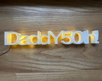 May include: A white illuminated sign that reads "Daddy 50th!" The letters are in a bold, sans-serif font, with a warm yellow glow emanating from within. The sign is plugged in and sits on a wooden surface.