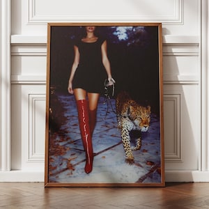 Könnte beinhalten: Gerahmter Kunstdruck mit einer Person in einem schwarzen Kleid und roten kniehohen Stiefeln, die einen Leoparden an der Leine führt. Der Leopard hat ein geflecktes Fell. Das Kunstwerk wird vor einer weißen Wand ausgestellt.