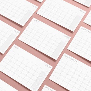 Puede incluir: Calendarios mensuales blancos de 2026 dispuestos sobre una superficie rosa. Cada calendario muestra un diseño de cuadrícula con el nombre del mes y el año impresos en la parte superior. Los calendarios están en ángulo, mostrando el diseño minimalista.