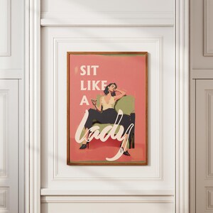 May include: A framed art print with the text "SIT LIKE A lady" in white script. The artwork depicts a woman in a yellow top and black pants, seated in a green chair, holding a drink. The background is a salmon pink.